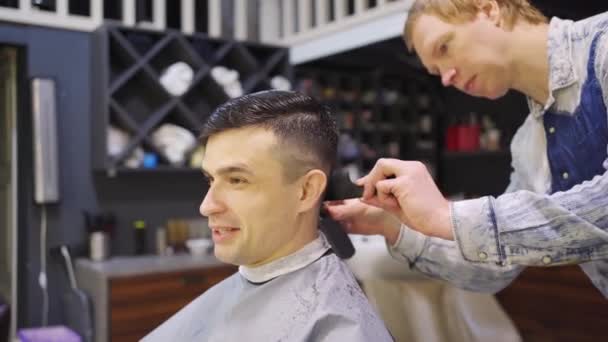 O cabeleireiro faz um corte de cabelo para um homem com um cortador de cabelo em uma barbearia — Vídeo de Stock