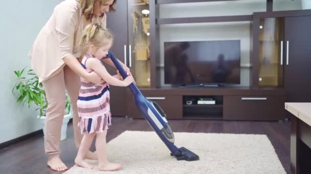 Мама учит маленькую дочь чистить ковер в комнате с пылесосом — стоковое видео