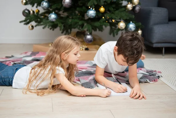 Children - a boy and a girl write letters to Santa Claus near the Christmas tree in the living room