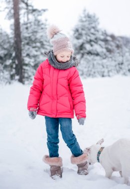 Parlak ceketli küçük kız kışın karlı ormanda köpeği Jack Russell Terrier ile oynuyor.