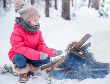 Parlak ceketli küçük bir kız kışın karlı ormanda oynar. Kız ateşin yanında ısınıyor..