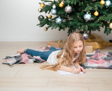 Küçük kız oturma odasındaki Noel ağacının yanında Noel Baba 'ya mektup yazıyor.