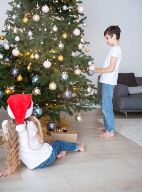 Çocuklar - bir oğlan ve bir kız Noel ağacının yanında oynuyorlar. Oturma odasının içi Noel ağacı ve süslemelerle dolu. Yeni yıl. Hediye vermek.