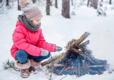 Parlak ceketli küçük bir kız kışın karlı ormanda oynar. Kız ateşin yanında ısınıyor..