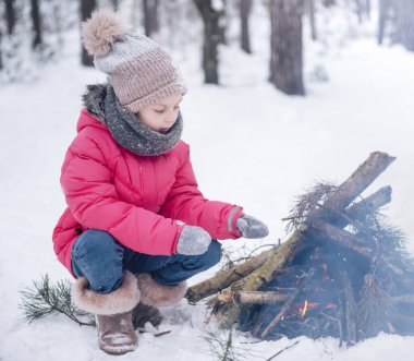 Parlak ceketli küçük bir kız kışın karlı ormanda oynar. Kız ateşin yanında ısınıyor..