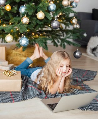 Küçük kız elinde dizüstü bilgisayarla Noel ağacının yanında yatıyor. Büyük bir oturma odasında Noel ağacı.