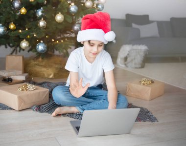 Bir çocuk elinde dizüstü bilgisayarla Noel ağacının yanında oturuyor. Büyük bir oturma odasında Noel ağacı.