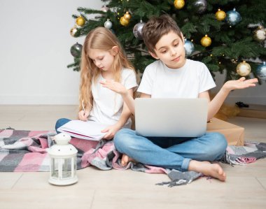 Büyük bir oturma odasında Noel ağacı. Çocuklar Noel ağacının yanında oynuyorlar. Dizüstü bilgisayarı olan bir çocuk arkadaşlarıyla iletişim kurar..