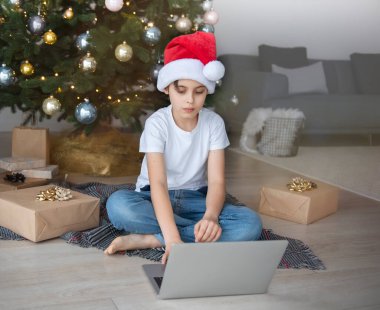 Bir çocuk elinde dizüstü bilgisayarla Noel ağacının yanında oturuyor. Büyük bir oturma odasında Noel ağacı.