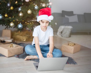 Bir çocuk elinde dizüstü bilgisayarla Noel ağacının yanında oturuyor. Büyük bir oturma odasında Noel ağacı.
