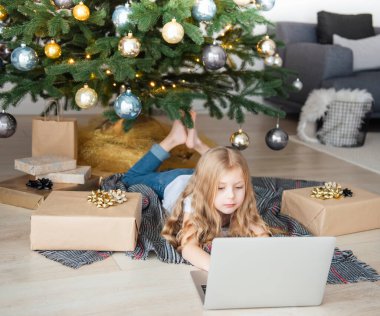Küçük kız elinde dizüstü bilgisayarla Noel ağacının yanında yatıyor. Büyük bir oturma odasında Noel ağacı.