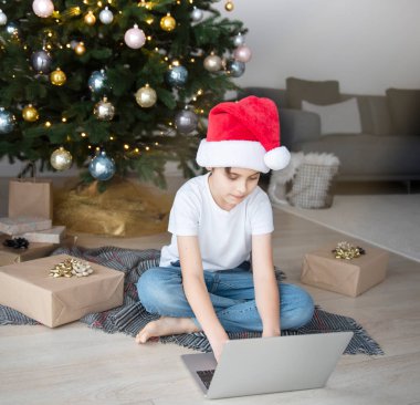 Bir çocuk elinde dizüstü bilgisayarla Noel ağacının yanında oturuyor. Büyük bir oturma odasında Noel ağacı.