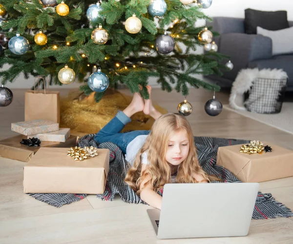 Küçük kız elinde dizüstü bilgisayarla Noel ağacının yanında yatıyor. Büyük bir oturma odasında Noel ağacı.