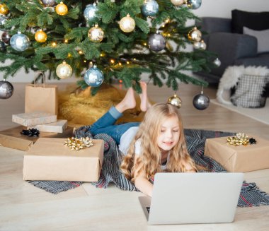Küçük kız elinde dizüstü bilgisayarla Noel ağacının yanında yatıyor. Büyük bir oturma odasında Noel ağacı.