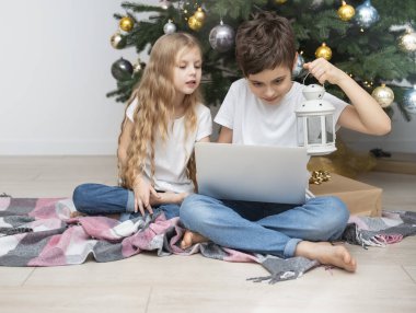 Büyük bir oturma odasında Noel ağacı. Çocuklar Noel ağacının yanında oynuyorlar. Dizüstü bilgisayarı olan bir çocuk arkadaşlarıyla iletişim kurar..