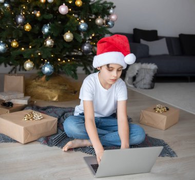 Bir çocuk elinde dizüstü bilgisayarla Noel ağacının yanında oturuyor. Büyük bir oturma odasında Noel ağacı.