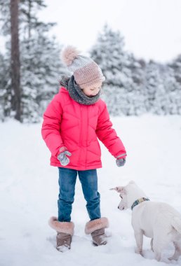 Parlak ceketli küçük kız kışın karlı ormanda köpeği Jack Russell Terrier ile oynuyor.