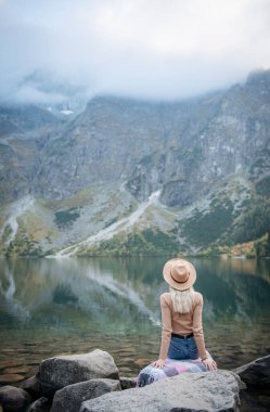 Genç bayan turist dağın zirvesinde ve güzel dağlarda oturuyor. Sportif kızla manzara, yeşil orman, tepeler, gökyüzü, göl. Seyahat ve turizm.