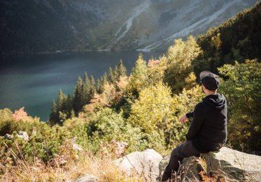 Bir tepenin üstünde dinlenen turist, dağların ve gölün muhteşem manzarasını izliyor..