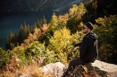 Bir tepenin üstünde dinlenen turist, dağların ve gölün muhteşem manzarasını izliyor..