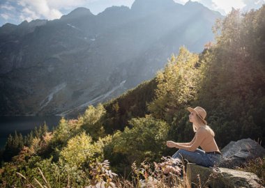Genç bayan turist dağın zirvesinde ve güzel dağlarda oturuyor. Sportif kızla manzara, yeşil orman, tepeler, gökyüzü, göl. Seyahat ve turizm.