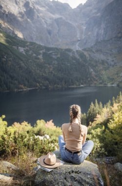 Genç bayan turist dağın zirvesinde ve güzel dağlarda oturuyor. Sportif kızla manzara, yeşil orman, tepeler, gökyüzü, göl. Seyahat ve turizm.
