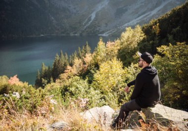 Bir tepenin üstünde dinlenen turist, dağların ve gölün muhteşem manzarasını izliyor..