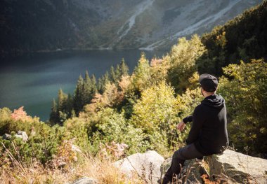 Bir tepenin üstünde dinlenen turist, dağların ve gölün muhteşem manzarasını izliyor..