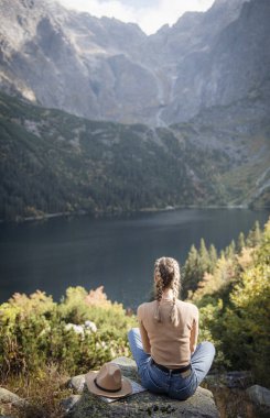 Genç bayan turist dağın zirvesinde ve güzel dağlarda oturuyor. Sportif kızla manzara, yeşil orman, tepeler, gökyüzü, göl. Seyahat ve turizm.