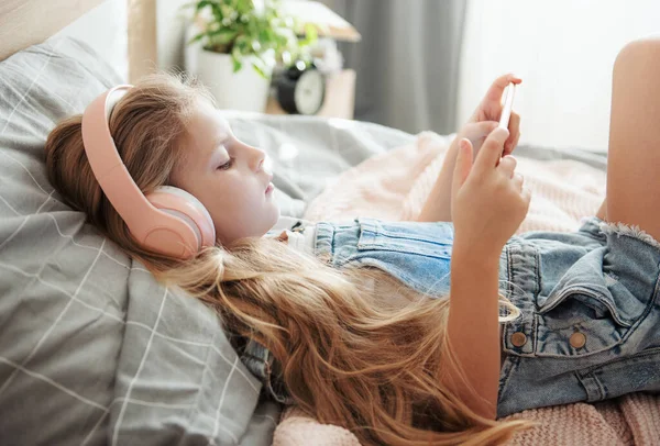 小女孩躺在床上一边听音乐一边拿着手机 — 图库照片