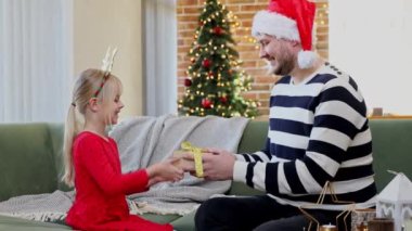 Noel baba şapkalı, şapkalı, Noel 'e şapkalı, kırmızı elbise giymiş kızına altın kurdeleli hediye paketi veren. Mutlu kız sarılması ya da hediye kutusu veren bir adama sarılması. Yüksek kalite 4k görüntü