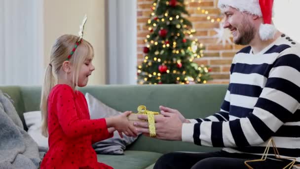 サンタの帽子の父は キャップはお祝いの赤いドレスを着た彼の娘に金リボンで紙のクリスマス 新年プレゼントのギフトボックスを与えます 幸せな女の子抱擁や男を抱擁 高品質4K映像 — ストック動画