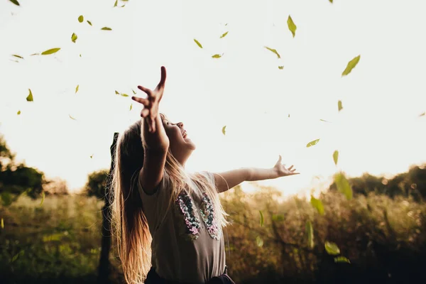 Το ξανθό κοριτσάκι παίζει με τα πράσινα φύλλα στον κήπο, χαμογελάει, διασκεδάζει, γελάει — Φωτογραφία Αρχείου