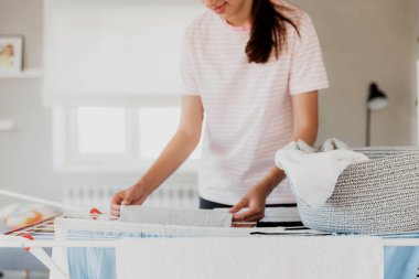 Happy young brunette caucasian woman smiling, hanging clean wet clothes laundry on drying rack at home. Female housewife hands closeup spreading laundry from basket. Girl puts towels after drying them