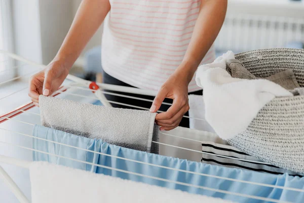 Woman hanging clean wet clothes laundry on drying rack at home laundry room. Detail of female housewife hands closeup holding, spreading and taking laundry basket in front of clothing rack dryer