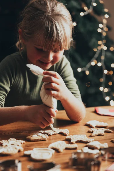 Christmas, New Year food preparation. Xmas gingerbread cooking, decorating freshly baked cookies with icing and mastic. Cute little girl decorate cookie on wooden messy table. DIY, creativity concept