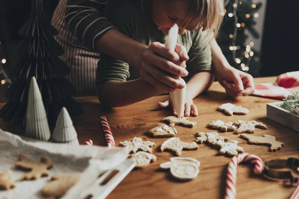 Christmas and New Year food preparation. Xmas gingerbread cooking and decorating freshly baked cookies with icing and mastic. Mom helping cute little daughter to decorate cookie on wooden messy table