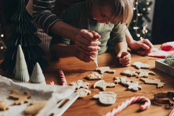 Christmas and New Year food preparation. Xmas gingerbread cooking and decorating freshly baked cookies with icing and mastic. Mom helping cute little daughter to decorate cookie on wooden messy table