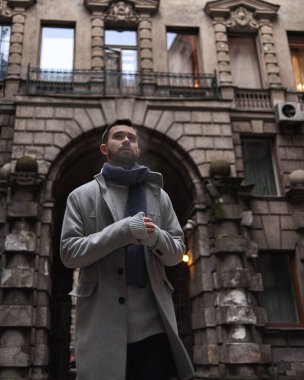 Gri paltolu ve mavi eşarplı sakallı bir adam tarihi bir evin kemerinde duruyor. Arka planda binanın eski ve güzel bir yüzü var..