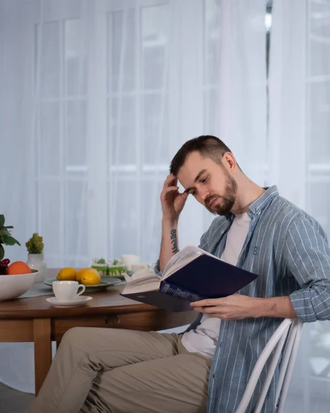 Mavi gömlekli ve bej pantolonlu sakallı bir adam masaya oturur ve kitap okur.