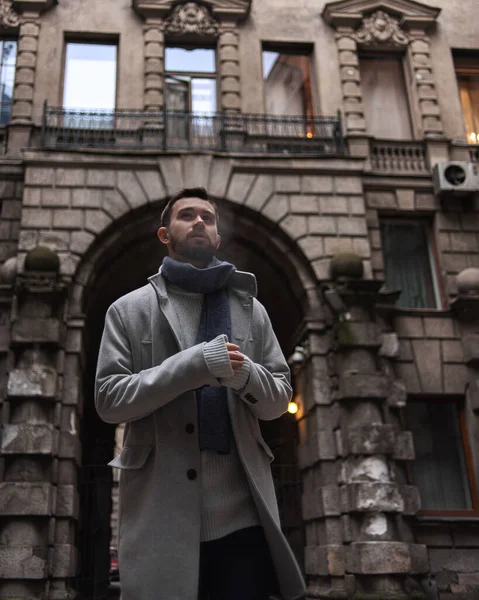 Gri paltolu ve mavi eşarplı sakallı bir adam tarihi bir evin kemerinde duruyor. Arka planda binanın eski ve güzel bir yüzü var..
