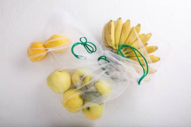 Muzlar, elmalar ve limonlar tekstil market poşetlerinde. Tekrar kullanılabilir çevre dostu ambalajlı sarı meyve ve sebzeler beyaz arka planda. Sıfır atık alışverişi, plastiksiz, kirliliği durdurun, ekolojik kavram. 