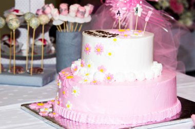 Çocuklar için pembe beyaz doğum günü pastası. Masada süslemeler var. Arka planda çeşitli kekler