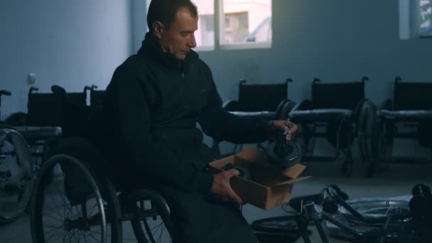 Πλευρική άποψη ενός αμερικανού εργάτη σε εργαστήριο σε εργοστάσιο κατασκευής αναπηρικών αμαξιδίων, που κάθεται σε πάγκο εργασίας χρησιμοποιώντας εργαλεία χειρός και συναρμολογώντας μέρη ενός προϊόντος, που κάθεται σε αναπηρικά αμαξίδια — Αρχείο Βίντεο