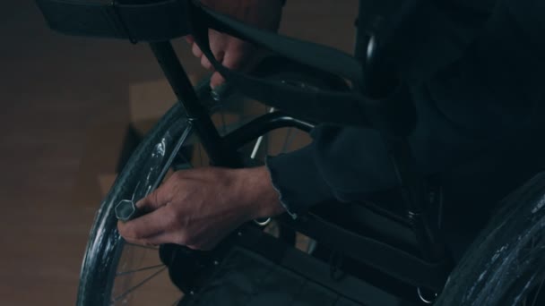 कारखान्यात व्हीलचेअर बनवण्याच्या कार्यशाळेत एका अमेरिकन पुरुष कामगाराचे साइड दृश्य, हाताची साधने वापरुन वर्कबेंचवर बसून आणि व्हील चेअरमध्ये बसून उत्पादनाचे भाग एकत्रित करते — स्टॉक व्हिडिओ