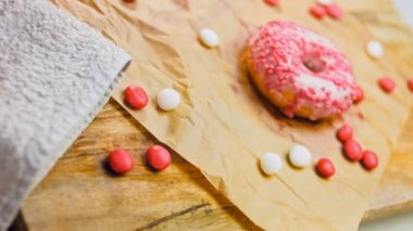 Pembe kremayla süslenmiş pembe çörekler. Çörekler şekerle süslenmiş bir kağıt parçasının üzerinde. Makro ve kaydırma atışı. Şekerler ağır çekimde hareket ediyor. Fırın ve yemek konsepti. Çeşitli renkli çörekler.