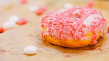 Pembe kremayla süslenmiş pembe çörekler. Çörekler şekerle süslenmiş bir kağıt parçasının üzerinde. Makro ve kaydırma atışı. Şekerler ağır çekimde hareket ediyor. Fırın ve yemek konsepti. Çeşitli renkli çörekler.