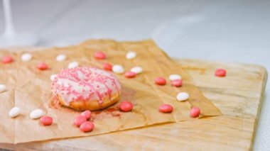 Pembe kremayla süslenmiş pembe çörekler. Çörekler şekerle süslenmiş bir kağıt parçasının üzerinde. Makro ve kaydırma atışı. Şekerler ağır çekimde hareket ediyor. Fırın ve yemek konsepti. Çeşitli renkli çörekler.