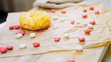 Sarı kremayla süslenmiş pembe çörekler. Çörekler şekerle süslenmiş bir kağıt parçasının üzerinde. Makro ve kaydırma atışı. Şekerler ağır çekimde hareket eder..