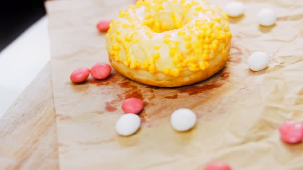 粉红色的甜甜圈装饰着黄色的糖霜 甜甜圈在用糖果装饰的一张纸上 宏观和滑翔机射击 糖果是慢动作的 — 图库视频影像